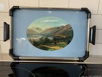 Top view showing the full tray with black and chrome edges, sky blue interior, and an oval hand-painted landscape image under glass.