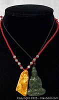Front view of green jade Buddha pendant on black cord with red bead and yellow ornamental stone pendant on red cord with beads.
