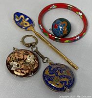 Set of cloisonne items arranged on white surface including spoon, bangle, keychain, pendant and blue fish charm.