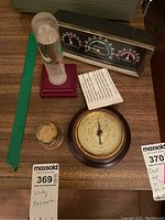 Four weather-related items on wooden table including a round wood framed barometer, sand art in a clear dome, storm glass on red base, and Airguide barometer with multiple gauges.