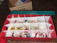 A red box with multiple individual cardboard compartments each holding a glass Christmas ornament including gold, silver, and painted white balls.