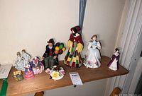 Photo of 9 porcelain figurines on wooden shelf including women in period dresses, a man holding colorful balloons, and floral decoration.