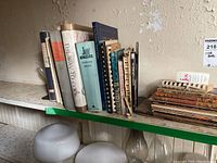 Photo shows a row of vintage cookbooks standing upright on a white shelf. Titles include The Way to Cook, Joy of Cooking, and other assorted old recipe books, all with worn and aged covers.