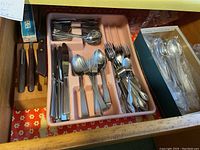 Drawer view showing various knives with wooden handles, flatware set in pink organizer tray, and box of additional spoons.