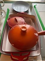 Overview of orange Le Creuset lidded pots stacked inside a Dansk rectangular red-handled baking pan on a counter.