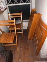 Photo showing six wooden folding chairs stacked and folded against a wall on a wooden floor.