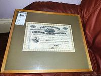 Framed stock certificate from Gold Mining Company of California displayed on a red couch, showing certificate with tan border and engraved miner images.