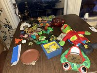 Photo showing the collection of various frog toys including the vintage Kermit the Frog plush, frog figurines, toy frogs, and decorative items arranged on a wooden table.