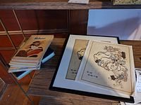 Two Japanese art prints, one framed and one unframed, displayed on shelf with stack of softcover books with Japanese art covers.