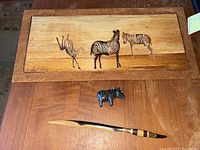 Frontal photo showing wooden wall art of three zebras, black zebra figurine, and wooden letter opener arranged on table.