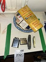 Group photo showing two vintage metal lighters, two folding pocket knives, and a dish holding matchbooks and a package of pipe cleaners.