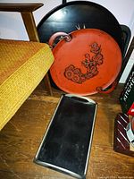 Photo showing four lacquered serving trays stacked, including a large black round tray with gold design, red floral embossed tray with handles, rectangular black tray with silver edges, and part of a large round black tray with handles.