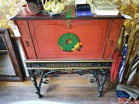 Front view of vintage drop front secretary desk showing red front panel with bird and flower painting, black legs with gold carvings, and ornate scrollwork.