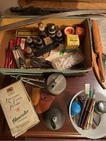 Box containing multiple vintage pencil sharpeners, several ink bottles including Skrip ink, Flexicolor pencil box, and various small accessories like a metal sharpener, compass, and pens.