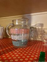 Full view of vintage glass pitcher with pink, white, and blue geometric band patterns, surrounded by clear drinking glasses and stemmed glassware on a red floral surface.
