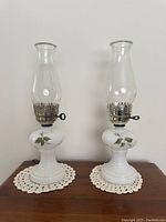 Pair of white milk glass hurricane lamps with clear chimneys and hand painted green branch and leaf appliques, each with metal wick holders.