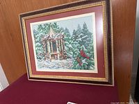 Front view of framed needlepoint artwork showing winter gazebo, pine trees and cardinals in detailed stitching with gold textured frame and maroon matting.