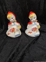 Pair of ceramic Little Red Riding Hood salt and pepper shakers side by side on black background showing full front view with floral dress and red hoods.