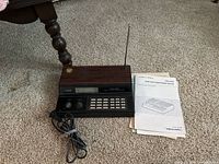 Photo showing radio scanner unit with woodgrain top, LCD display, numeric keypad, knobs, power cord, and user manual on carpeted floor.
