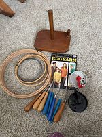 Photo showing four wooden embroidery hoops, seven rug hooks with wooden and blue handles, two yarn winders (one white and one black), and the vintage 'Rug Yarn' booklet.
