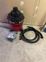 Shop Vac 12 gallon wet dry vacuum with two hoses laying on floor beside it and a sealed cloth filter bag. Cobwebs and dust are visible on surfaces.