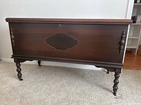 Full front view of the vintage wooden cedar chest showing the carved medallion design, turned spindle side accents, leg design, and wheels.