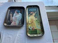 Two Coca Cola metal trays shown together on white cloth surface under window light