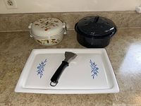 Vintage Corning broil plate P-35B with blue floral designs, metal scraper with black handle, Corning casserole with autumn leaf pattern, and Halls Superior Stoneware black speckled Dutch oven with lid placed on kitchen counter.