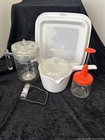 Photo showing all five vintage kitchen items on black cloth: Corning saucepan, clear Pyrex coffee pot, white microwave browner tray, nut chopper with orange plunger, and metal biscuit cutter.