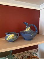 Two pieces of Roseville Pottery in Freesia pattern on wooden shelf with dark red wall background, showing both the Ewer and Cream Pitcher.