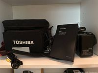 Photo of Toshiba branded carrying bag, VHS cassette adapter, power charger and AV cable arranged on shelf.
