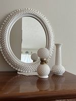 Full view of the three white hobnail items together: oval mirror, small oil lamp, tall bud vase on wooden surface