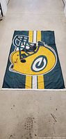 Photo of full size Green Bay Packers flag laid flat on floor showing football helmet and striped design.