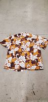 Full view of the short-sleeve shirt laid flat on a concrete floor showing the front design with orange, brown, and white floral pattern.