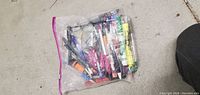 Clear plastic zipped bag containing over 25 assorted pens, markers, and highlighters on a concrete floor with a shoe partially visible.