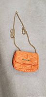 Orange quilted satchel purse with gold-tone chain strap shown against a concrete background