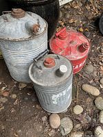 Photo of three antique metal gas cans on a ground surface with rocks and leaves showing different sizes and colors.