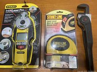 Photo of new Stanley IntelliLaser Pro laser level in sealed plastic packaging alongside STRAIT-LINE laser level in packaging, both in yellow and black color schemes.