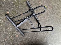 Black metal bike stand with support bars and wheel holder, resting on pavement outdoors.