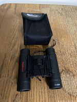 Tasco black binoculars positioned upright next to their black carrying case on a wooden surface.