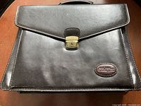 Front view of dark brown leather travel bag with flap and lock closure, Bugatti logo patch visible.