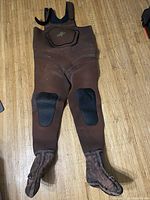 Full view of brown fishing chest waders showing black reinforced knee patches and attached boot section, laid flat on a wooden floor.