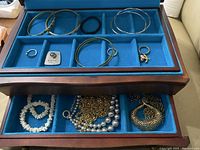 Wooden jewelry box open showing blue felt lined compartments filled with multiple bracelets, necklaces including puka shell necklaces, rings, and a hair tie.