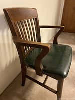 Solid wooden chair with slatted backrest and green seat cushion, wooden armrests, photographed from front-side angle.
