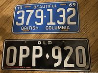 Photo showing two old license plates: a 1969 British Columbia plate with blue background and white text, and a Queensland plate with black background and white text.