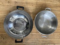 Top-down view of two stainless steel woks with black and silver handles, showing the interior surfaces and side handles.