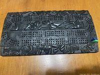 Top view of rectangular dark stone cribbage board with intricate carvings and multiple rows of peg holes. Four colored pegs visible at bottom right corner.