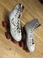 Pair of vintage white leather roller skates with red wheels on wooden floor, front angle showing laces and wheel condition.