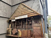 Front and side close-up of the metal wall hanging showing rusted details, door, windows, and sign.