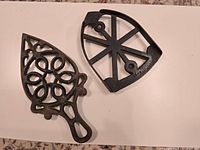 Photo showing two antique cast iron coasters featuring distinct decorative patterns on flat platforms designed to hold hot irons, with aged patina.
